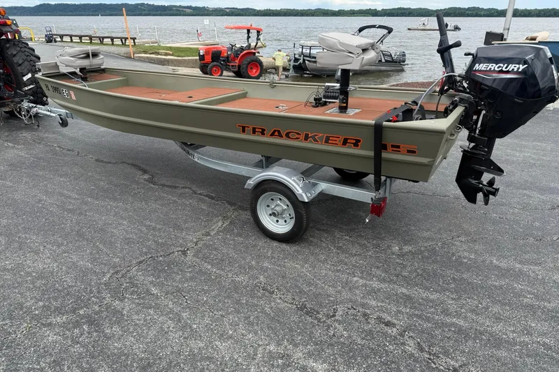 The Image of 2025 Tracker Grizzly 1542 boat on trailer with Mercury outboard motor by a lake. - 0