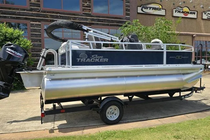 Slide: The Image of 2026 Sun Tracker Bass Buggy 16 XL Select pontoon boat on trailer, parked outdoors. - 2