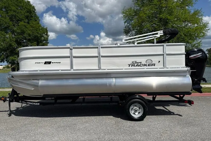 Slide: The Image of 2026 Sun Tracker Party Barge 18 DLX pontoon boat on trailer, parked by a lake. - 1
