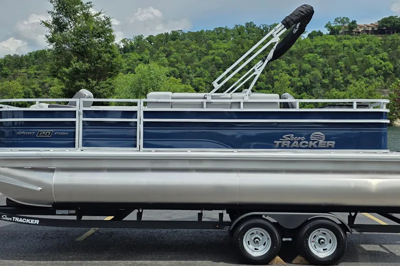 The Image of 2026 Sun Tracker SportFish 20 DLX pontoon boat on trailer, parked near lush greenery. - 0