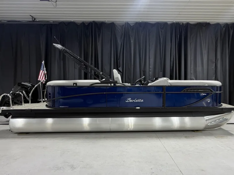 Slide: The Image of A 2027 Barletta Cabrio22QC pontoon boat is in a showroom. It features a blue hull and an American flag. - 1