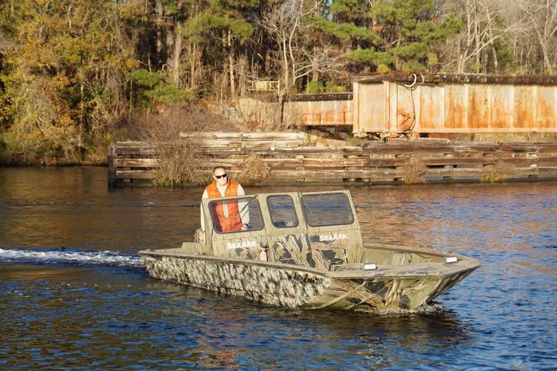 The Image of 2019 SeaArk Predator 220 AK SC boat navigating a calm river with wooded shoreline. - 1
