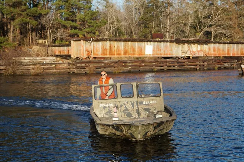 Slide: The Image of 2019 SeaArk Predator 220 AK SC boat navigating a calm river near a rustic dock. - 8