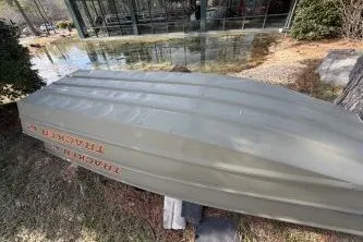 Slide: The Image of 2026 Tracker Grizzly 12 Jon boat upside down on grass near a glass building. - 2