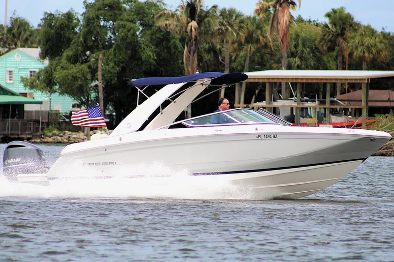 Slide: The Image of 2021 Regal 26 OBX boat cruising on water with American flag, lush greenery in background. - 44