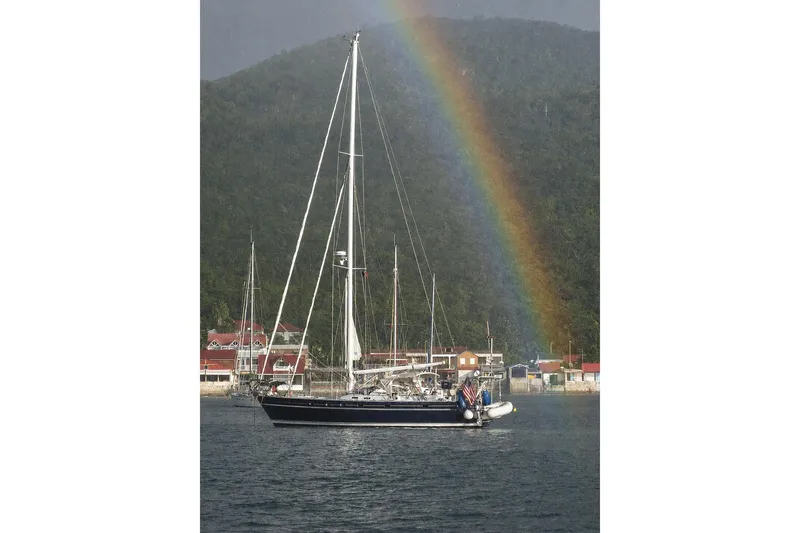 The Image of Sailboat Contest 46/48 (1996) under rainbow near forested hills and coastal village. - 0