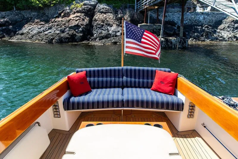 Slide: The Image of Hinckley Picnic Boat 36 Classic, 2000 model, with striped seating and American flag, near rocky shoreline. - 10