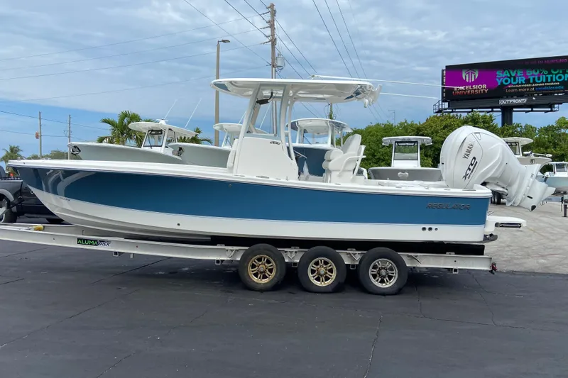 Slide: The Image of 2027 Regulator 26XO boat on trailer, blue and white design, parked outdoors. - 2