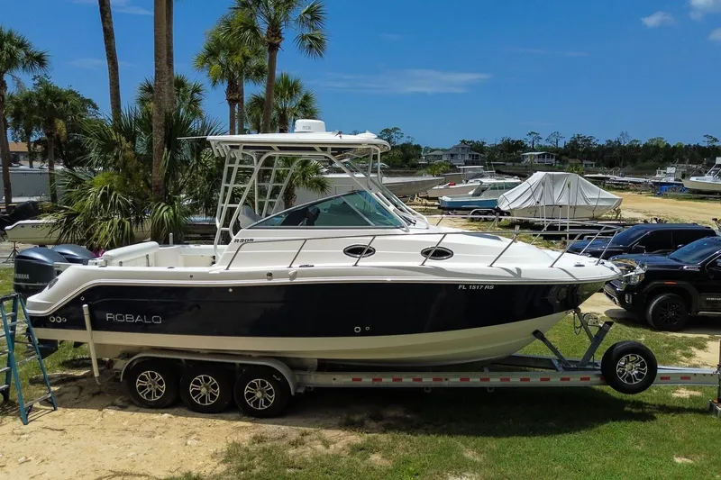 The Image of 2018 Robalo R305 Walkaround boat on trailer, surrounded by palm trees and parked vehicles. - 0