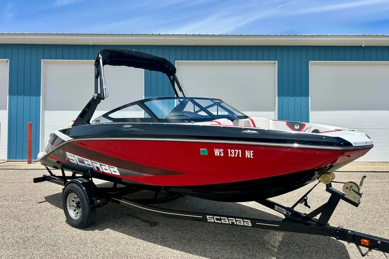 The Image of 2021 Scarab 195 ID boat in red and black, parked on a trailer outside a blue building. - 1