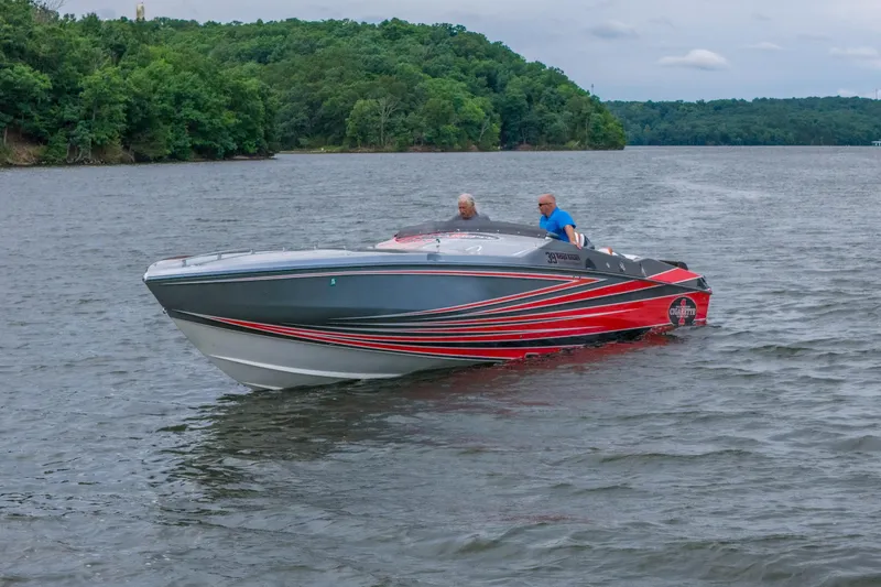 The Image of 2006 Cigarette 39' Top Gun boat cruising on a scenic lake with lush green shoreline. - 1