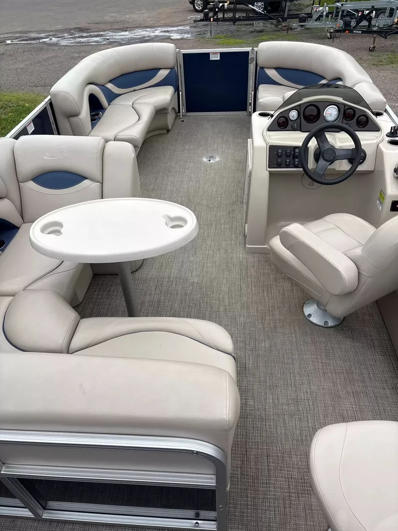 Slide: The Image of The image shows the interior of a 2016 Sylvan MIR 8520 CNF pontoon boat with beige seating and a table. - 6