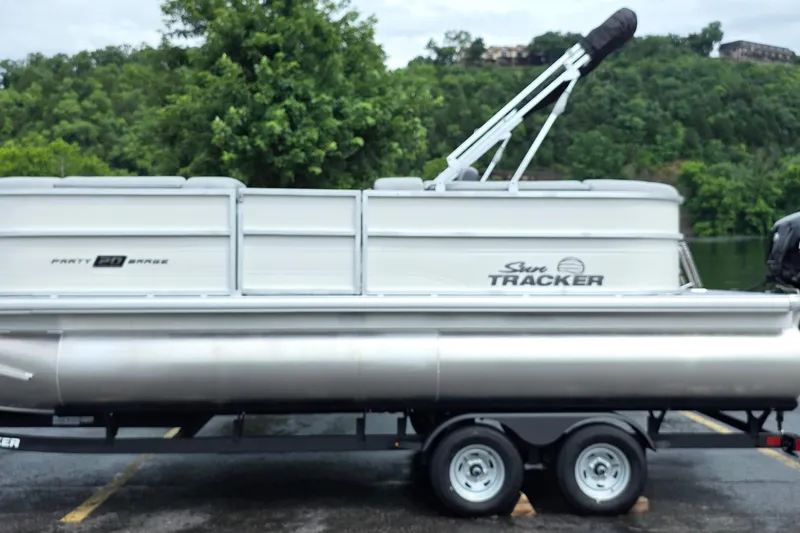 The Image of 2026 Sun Tracker Party Barge 20 DLX pontoon boat on trailer, parked near lush greenery. - 0