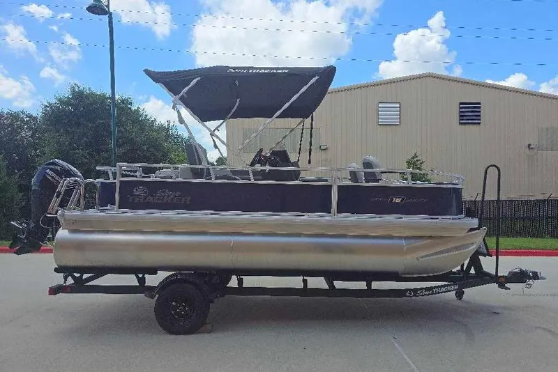 The Image of A Sun Tracker Bass Buggy 18 DLX pontoon boat on a trailer, with a black canopy and outboard motor. - 0