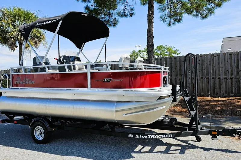 Slide: The Image of 2026 Sun Tracker Bass Buggy 16 XL Select pontoon boat on trailer, parked outdoors. - 2
