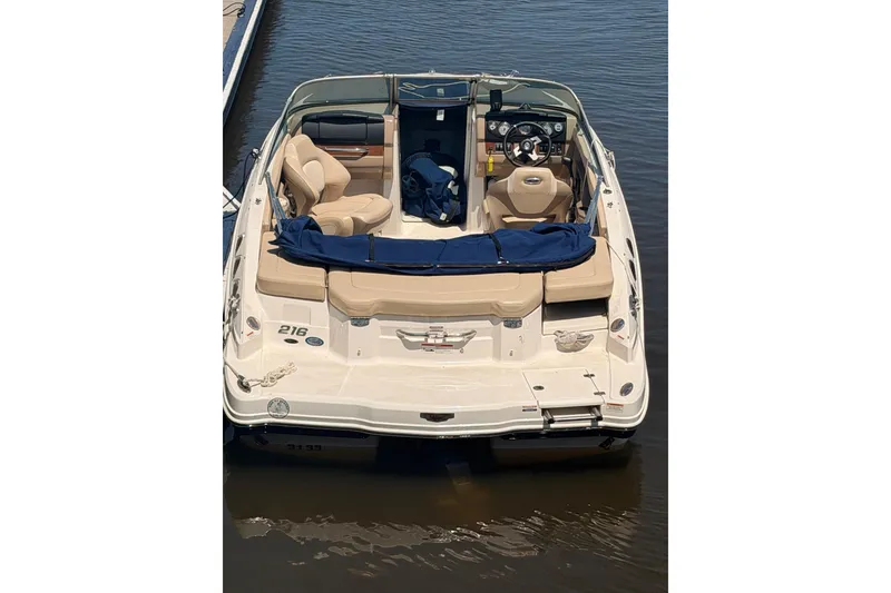 Slide: The Image of 2012 Chaparral 216 SSi boat docked on calm water, featuring beige seating and blue cover. - 2