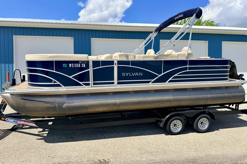 Slide: The Image of 2018 Sylvan 8522 Mirage Fish pontoon boat on trailer, parked near blue building. - 2