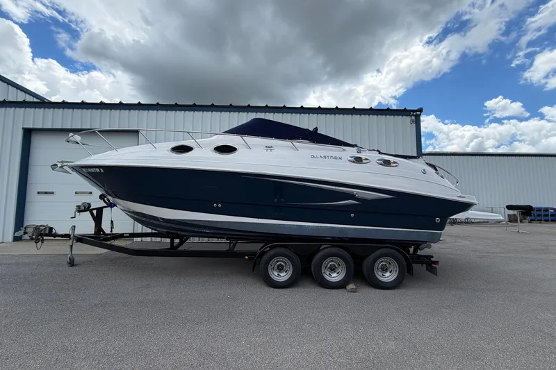 The Image of 2015 Glastron GS 289 boat on trailer, parked outside a warehouse under cloudy sky. - 1