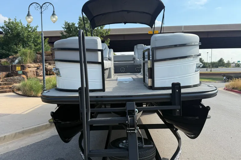 Slide: The Image of 2024 Regency 250 DL3 pontoon boat on trailer, parked outdoors near greenery and a bridge. - 4