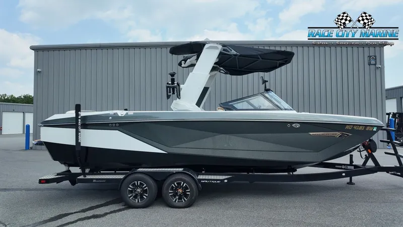 The Image of A 2022 Nautique G25 boat on a trailer in front of a gray building. - 1