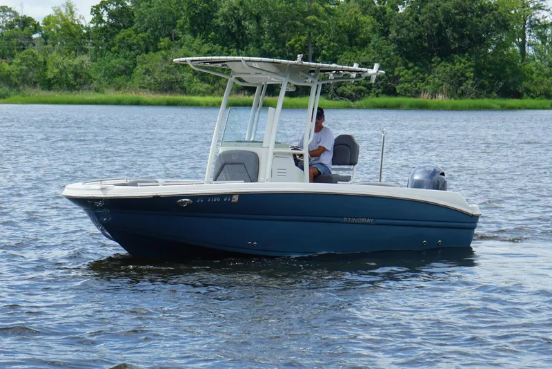 The Image of 2021 Stingray 216 CC boat cruising on a calm lake with lush green shoreline. - 0