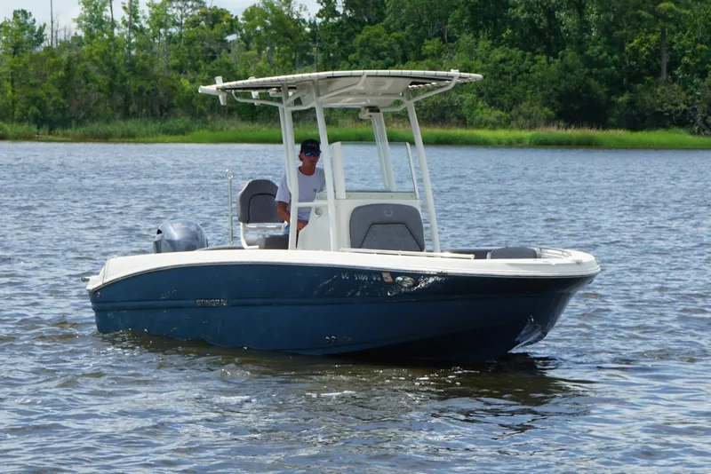 Slide: The Image of 2021 Stingray 216 CC boat cruising on a calm lake with lush green shoreline. - 15
