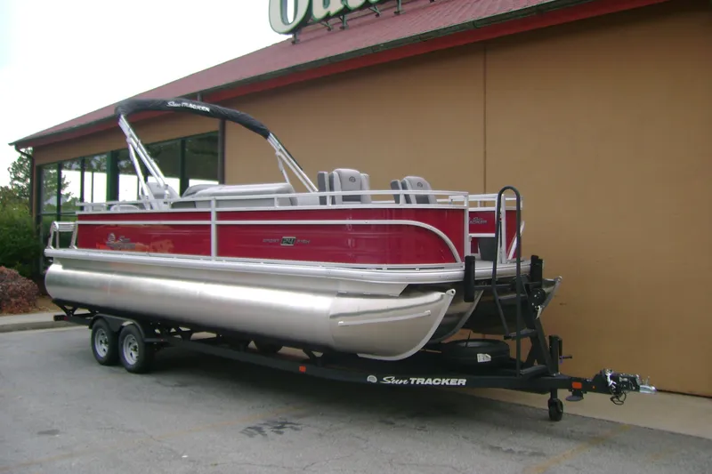 Slide: The Image of 2026 Sun Tracker Sportfish 24 XP3 pontoon boat on trailer, parked outside a building. - 2