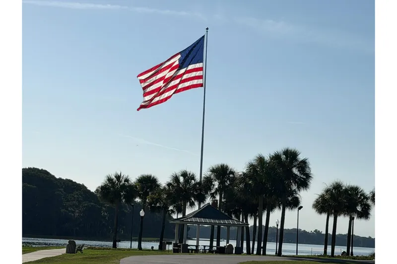 Slide: The Image of American flag waving near palm trees by a waterfront pavilion under a clear sky. - 66