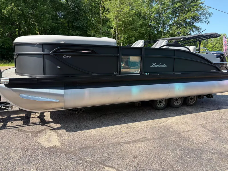 The Image of A 2027 Barletta Cabrio C22UC/TT pontoon boat on a trailer, surrounded by trees. - 1