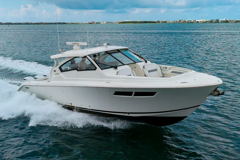 Slide: The Image of A 2021 Pursuit DC 365 Dual Console boat speeds across the water, surrounded by a calm coastal landscape. - 49
