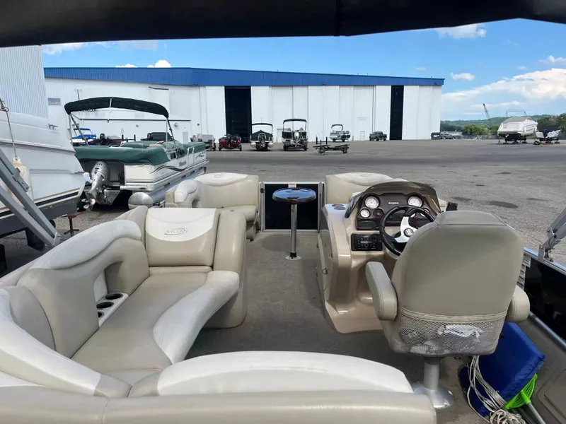 Slide: The Image of The image shows the interior of a 2015 Sylvan 8522 CNF pontoon boat. It features comfortable seating and a modern dashboard. Other boats and a large building are visible in the background. - 17