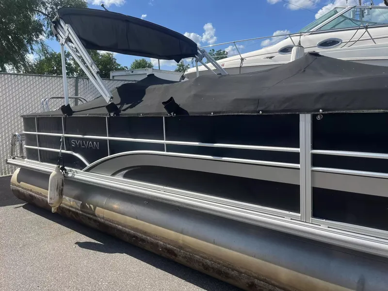 Slide: The Image of A 2015 Sylvan 8522 CNF pontoon boat with a black cover, parked on an asphalt lot. - 18
