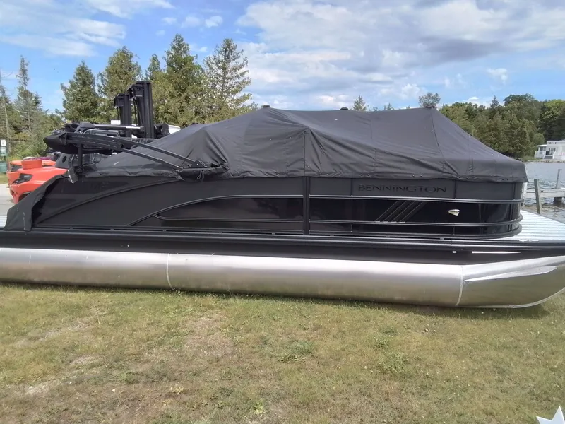 Slide: The Image of A 2026 Bennington 23M1SB pontoon boat sits on grass, covered with a black tarp. - 2