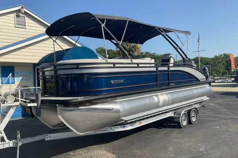 Slide: The Image of A 2025 Harris Solstice 250 SL pontoon boat on a trailer in front of a building. - 2