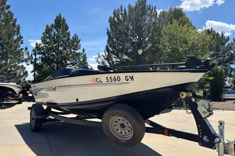 Slide: The Image of 2021 Alumacraft Classic 165 boat on trailer, parked outdoors with trees in background. - 2