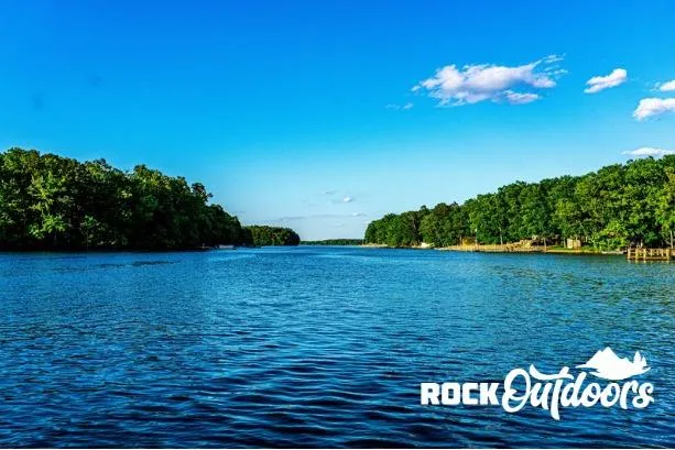 Slide: The Image of Scenic lake view with lush green trees under a clear blue sky, featuring Rock Outdoors logo. - 10