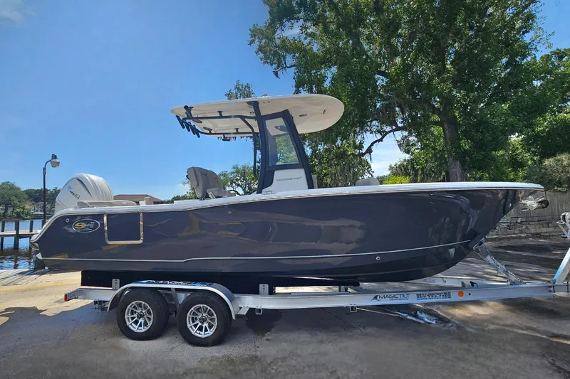 Slide: The Image of 2027 Sea Hunt Ultra 245 SE boat on trailer, parked near water, under clear blue sky. - 2
