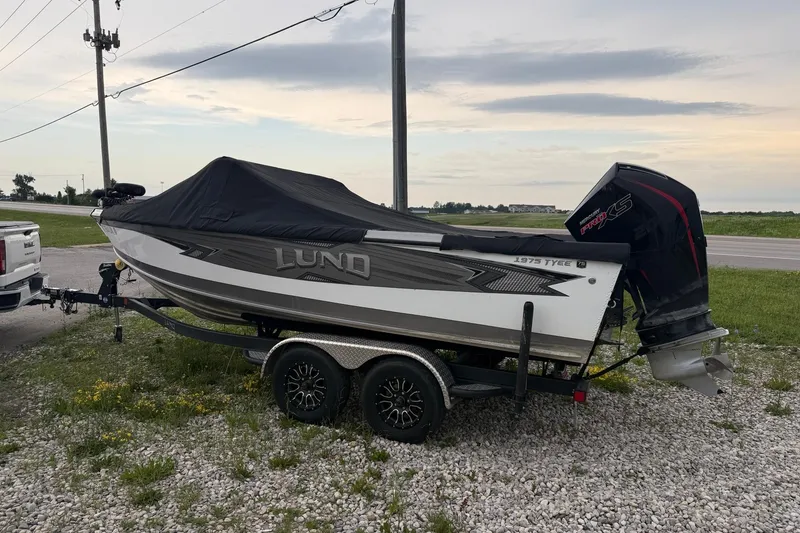 Slide: The Image of 2023 Lund 1975 Tyee boat on trailer, covered, with outboard motor, parked on gravel. - 1