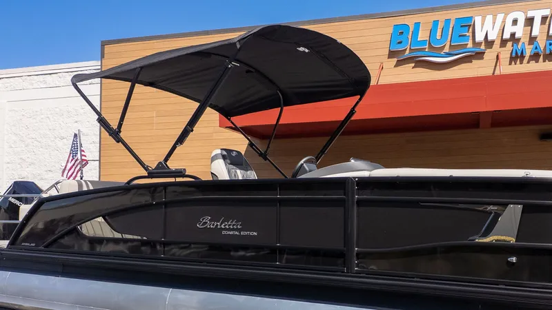 The Image of A Barletta Lusso 25UE pontoon boat with a black Bimini top, parked in front of a building. - 1