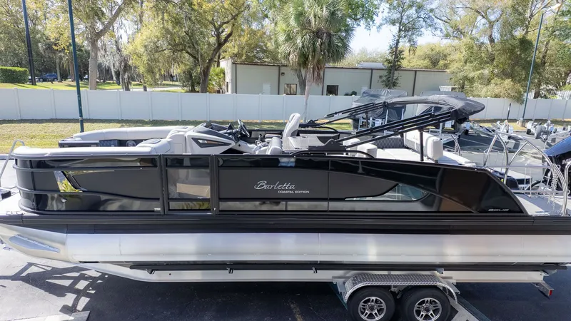 The Image of A sleek Barletta Lusso 25UE pontoon boat on a trailer, surrounded by trees. - 1