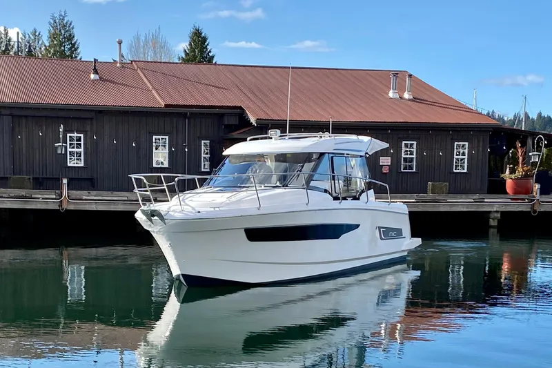 The Image of 2023 Jeanneau NC Weekender 895 boat docked by rustic building on calm water. - 0