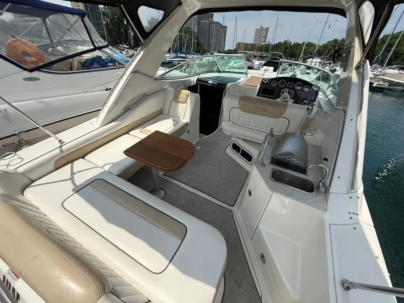 Slide: The Image of The image shows the cockpit of a 2010 Sea Ray Sundancer 310. It features white upholstery, a small wooden table, and a compact kitchenette. Other boats are visible in the harbor background. - 10