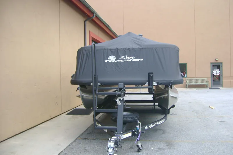 Slide: The Image of 2026 Sun Tracker Party Barge 20 DLX covered on trailer, parked near building. - 7