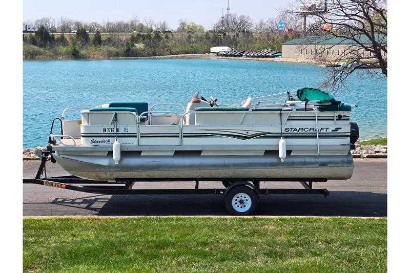 The Image of 1998 Starcraft pontoon boat on trailer by a lake, ready for transport. - 0