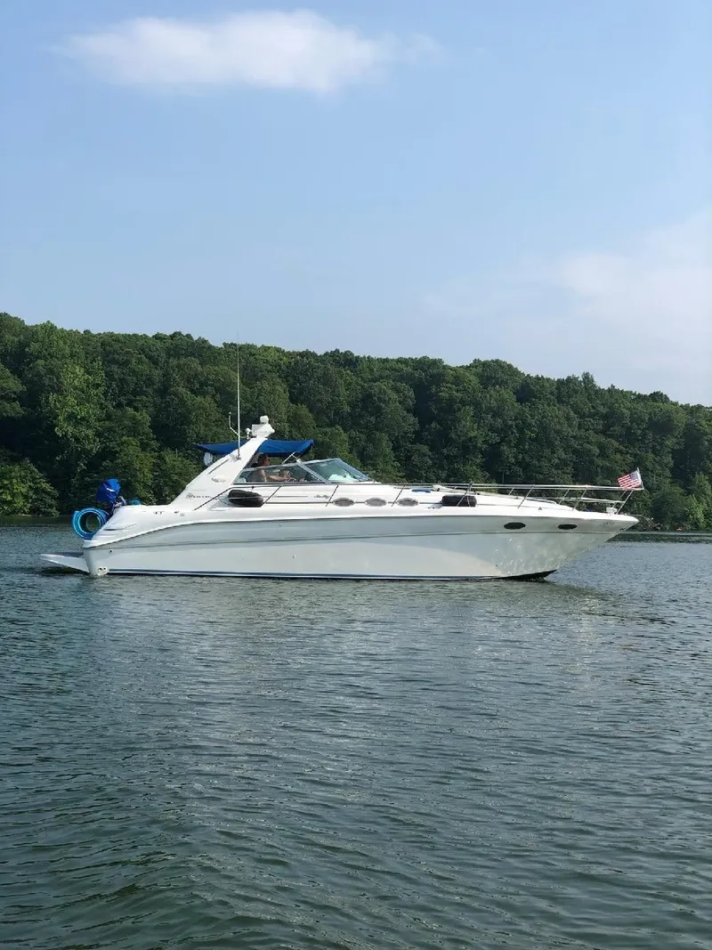 The Image of A white motorboat, the 1997 Sea Ray Sundancer 370, floats on calm water near a wooded shoreline. - 0