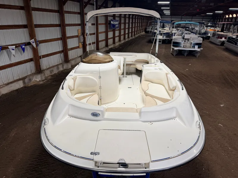 Slide: The Image of A 2003 Glastron DX215 deck boat in a warehouse, featuring beige interior and a Bimini top. - 10