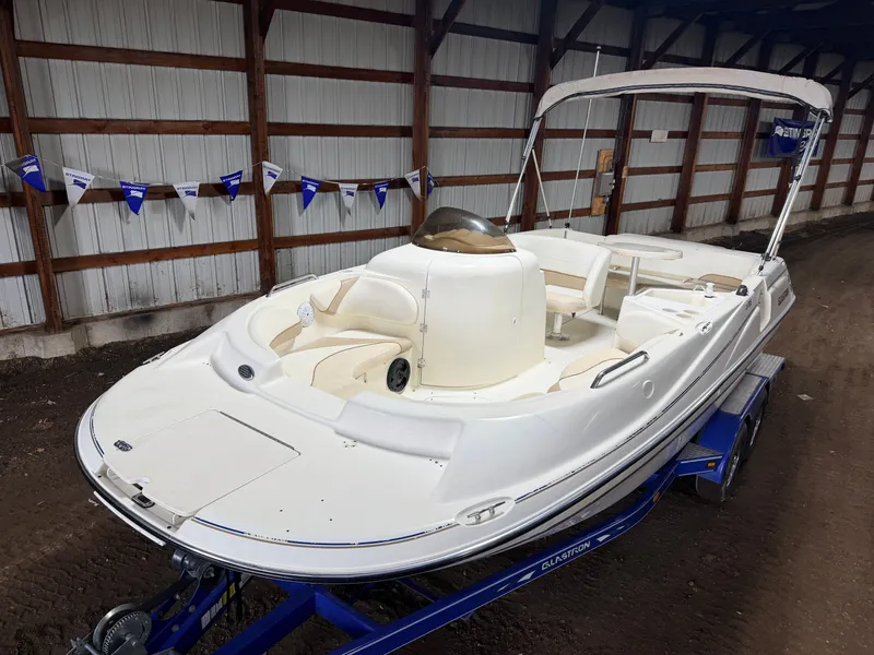 The Image of A white 2003 Glastron DX215 deck boat on a blue trailer inside a warehouse. - 1