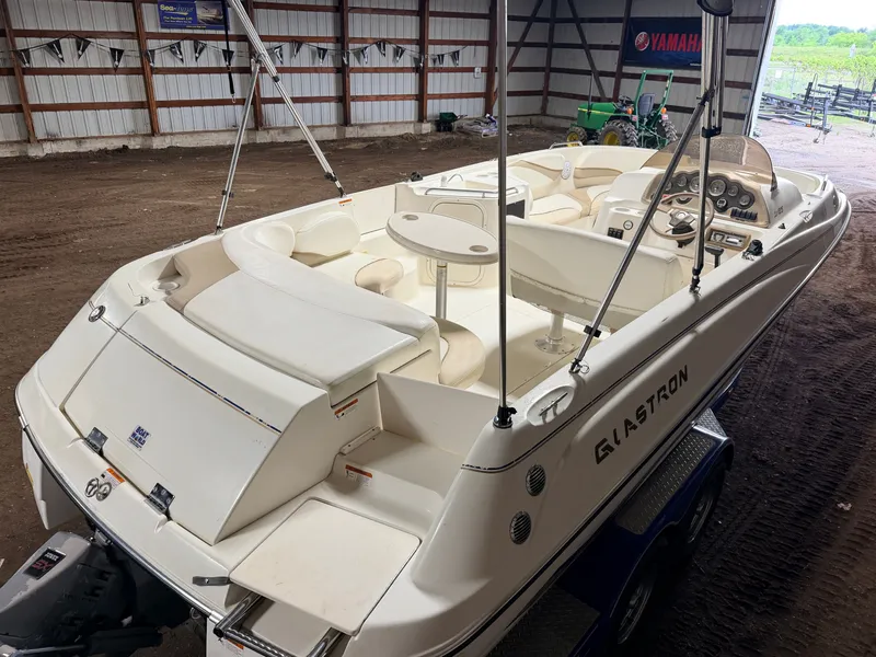 Slide: The Image of A 2003 Glastron DX215 deck boat is in a warehouse. It features white seating and a helm console. - 7