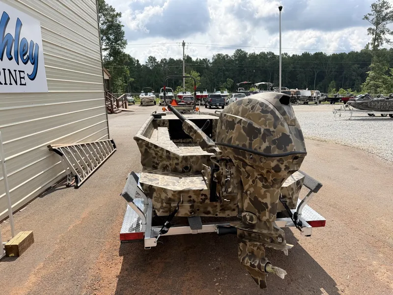 Slide: The Image of A camouflaged aluminum boat with an outboard motor on a trailer, surrounded by trees and buildings. - 2