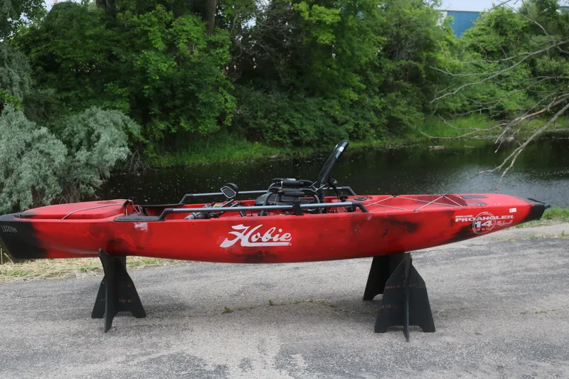 The Image of Red 2023 Hobie Mirage Pro Angler 14 kayak displayed outdoors near a serene pond. - 1